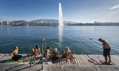 People in bathing costumes relaxing beside Lake Geneva, Switzerland, at the Bains des Paquis public bath, with the landmark fountain, known as Jet d'Eau, in the background.